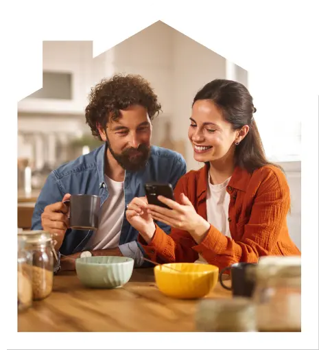 A man and woman sit at a kitchen table with mugs and bowls, both smiling and looking at a smartphone together.