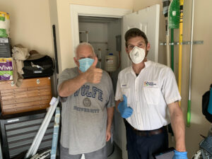 Two men wearing masks and gloves stand in a utility room; one gives a thumbs up while the other wears a work uniform and holds a tablet.