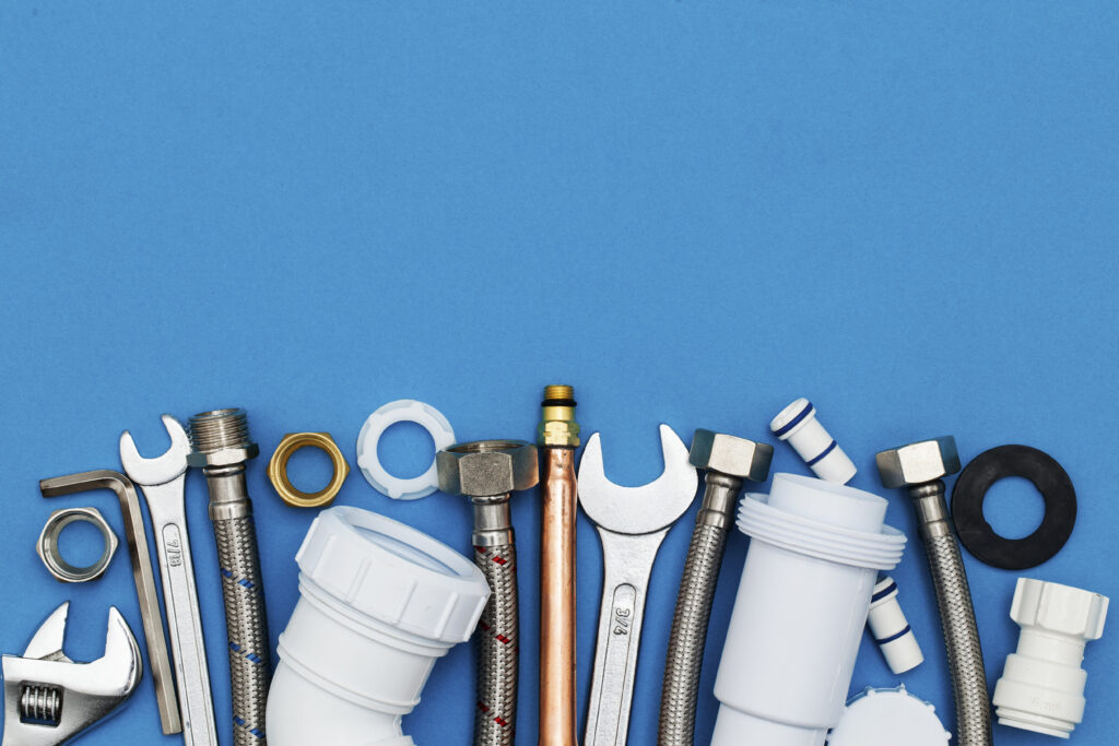 Assorted plumbing tools, metal hoses, fittings, wrenches, and plastic pipe parts arranged in a row on a blue background.