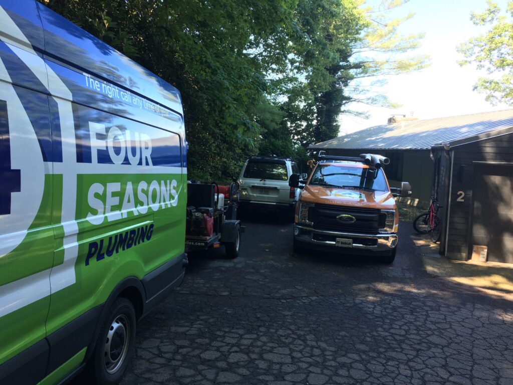 A Four Seasons Plumbing van and two trucks are parked on a cracked driveway next to a black building with a garage and a bicycle visible.