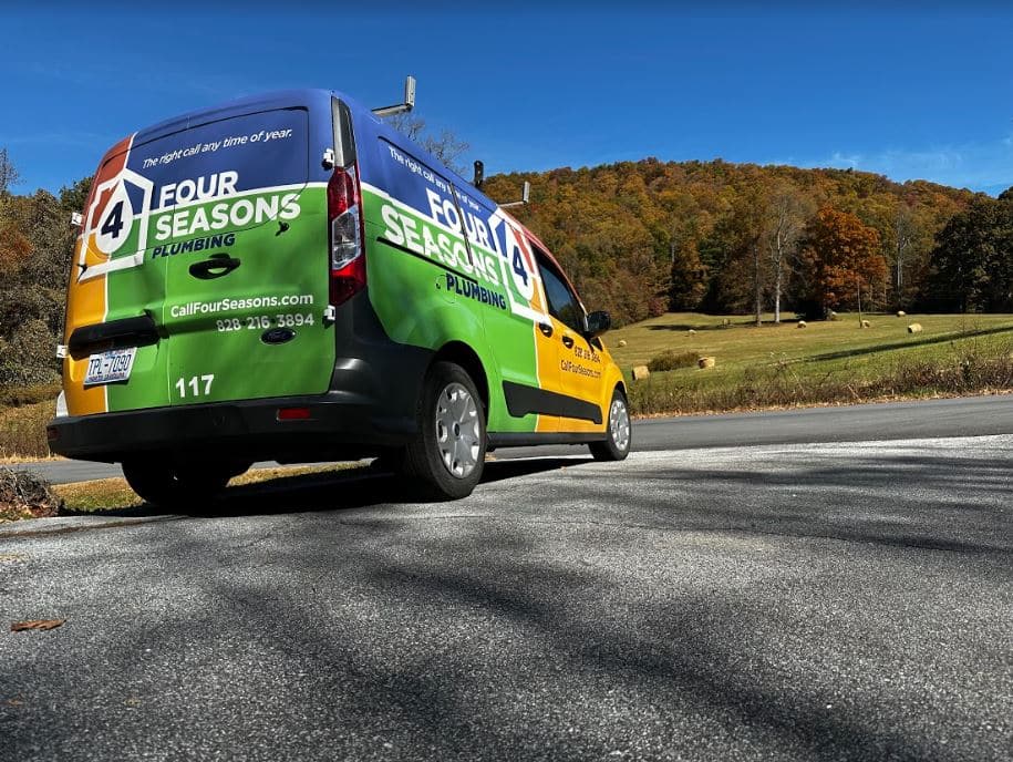 A Four Seasons Plumbing van is parked on a rural road beside a grassy field and trees with autumn foliage.