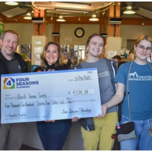 Four people stand indoors, smiling and holding a large donation check for $4,574.34 made out to the Asheville Humane Society from Four Seasons Plumbing, showcasing their strong commitment to community support.
