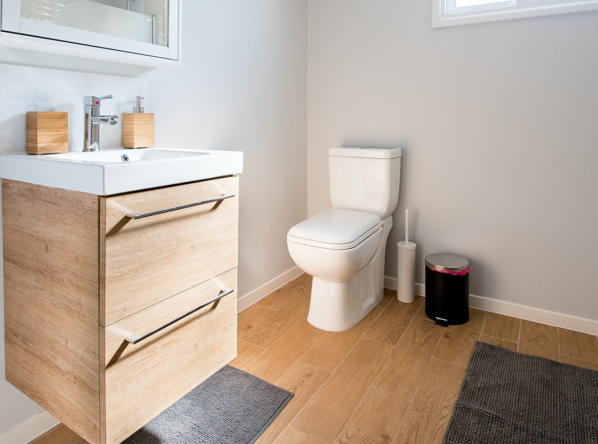 A modern bathroom with a white toilet, wooden vanity with sink, two grey floor mats, a small black trash bin, and a toilet brush on a wooden floor.