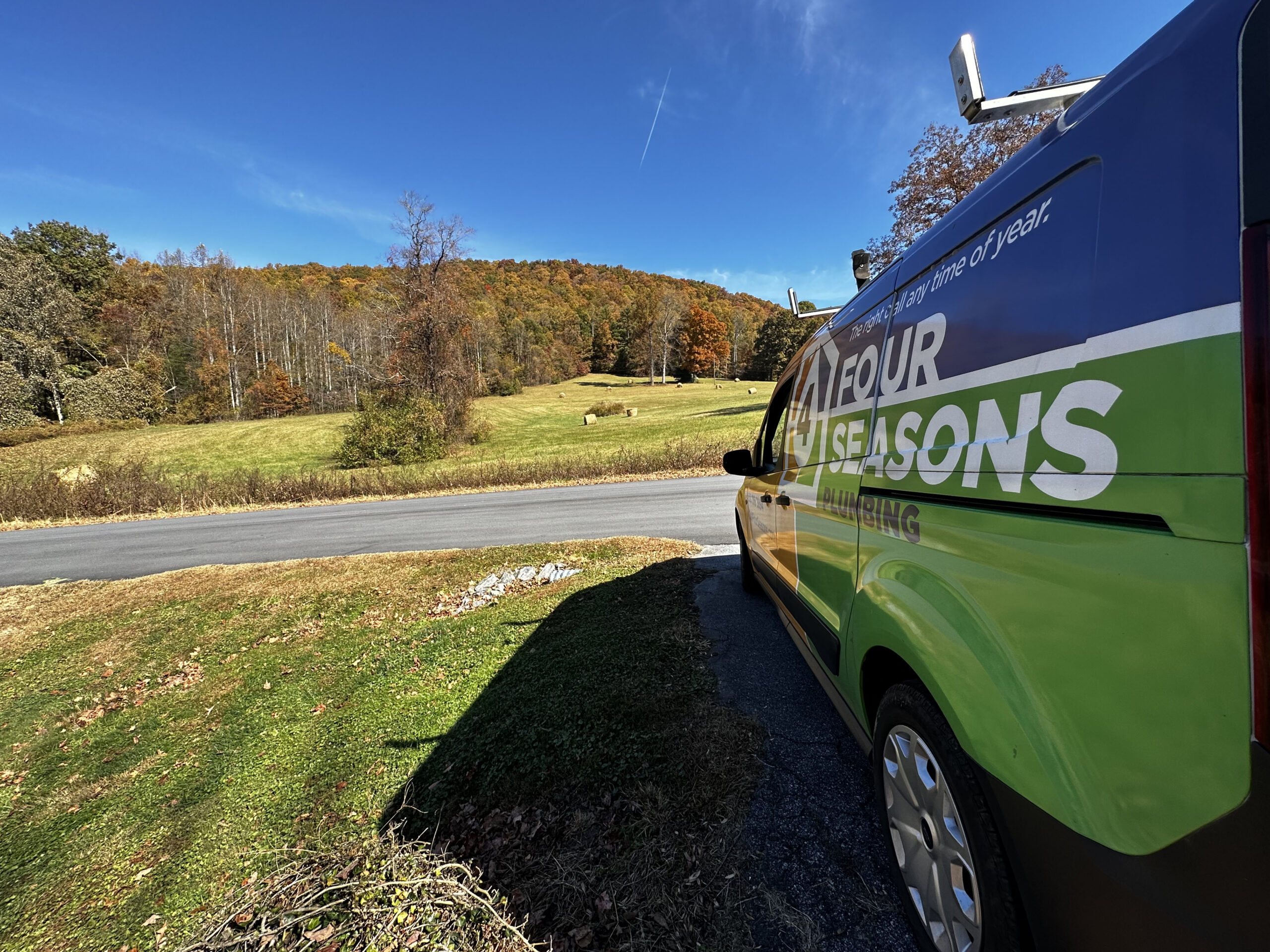 A Four Seasons van is parked near a road, facing a grassy field with trees and hills in the background under a clear blue sky.