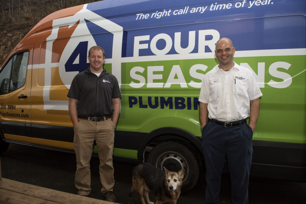 Two men and a dog stand in front of a Four Seasons Plumbing van with a colorful logo and slogan, “The right call any time of year.”.