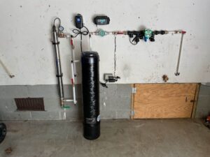 %Title% | Four Seasons Plumbing Water filtration and treatment system mounted on a wall, featuring pipes, a tall black filter tank, electronic control units, and a wooden hatch nearby.