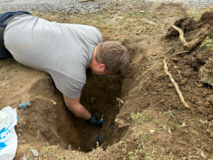 %Title% | Four Seasons Plumbing A person in a gray shirt is kneeling and working on pipes in a hole dug in the ground, with exposed soil and roots visible.