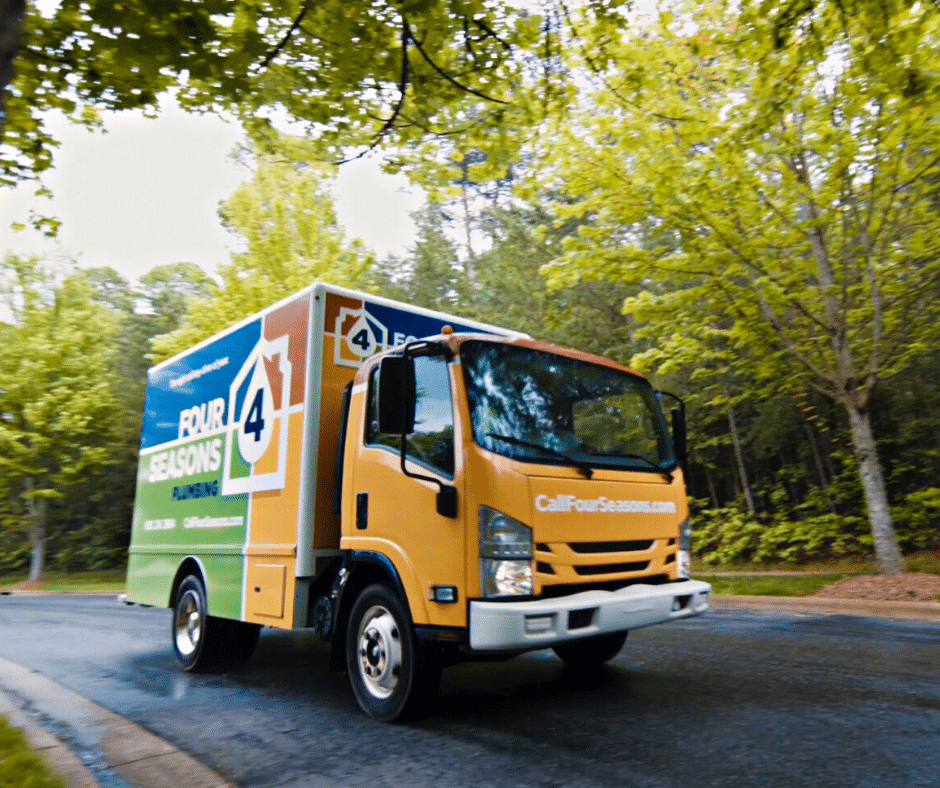 A Four Seasons Plumbing service truck drives on a wet road through a leafy, green neighborhood.