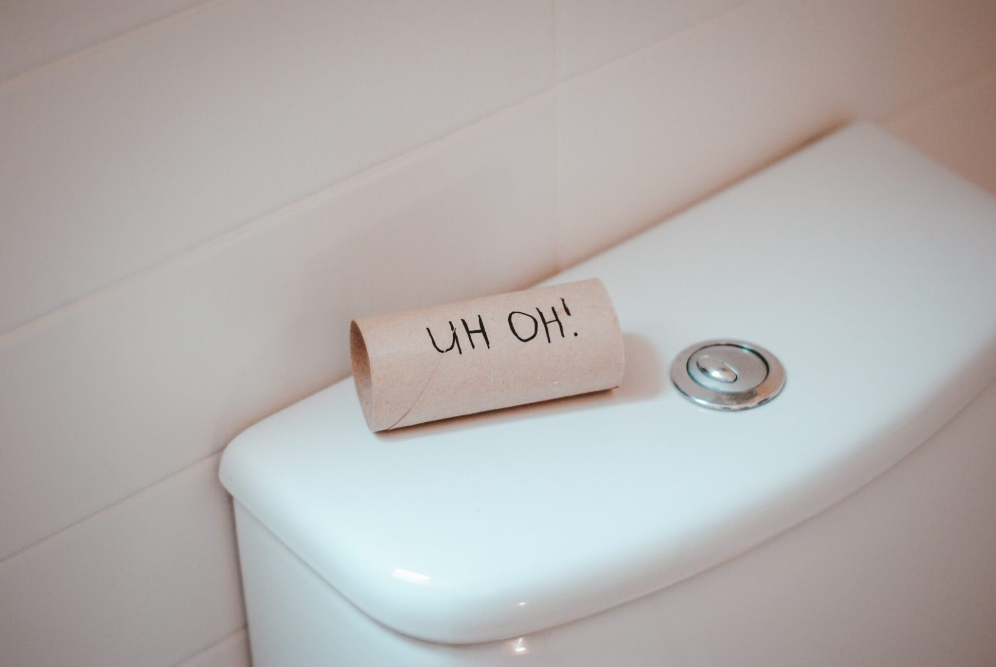 An empty toilet paper roll labeled "UH OH!" sits on top of a white toilet tank.
