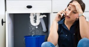 %Title% | Four Seasons Plumbing A woman sits on the floor, looking concerned while talking on the phone. Water leaks from a pipe under the sink into a blue bucket.