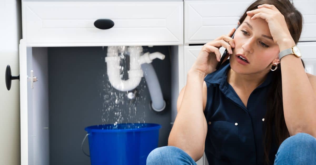 A woman sits on the floor, looking stressed while calling for help as water leaks from a pipe under a sink into a blue bucket, worried her water bill is high.