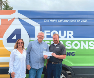 Three people stand in front of a Four Seasons Heating van; the man in the middle is handing a check to the man on the right, showcasing their commitment to supporting the local community.