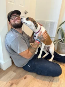 A man sitting on the floor smiles as a brown and white dog with a pink collar licks his face, capturing a joyful moment of community and companionship.