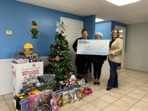 Three women stand beside a decorated Christmas tree, holding a large check for Toys for Tots; the festive scene highlights community spirit with assorted toys displayed on the floor and in donation boxes nearby.